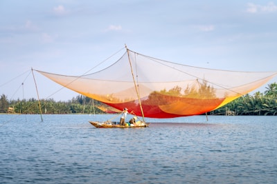 A fisher stands in a small boat on a calm body of water, operating a large, colorful fishing net suspended by poles. The net has a gradient of orange and yellow hues and stretches across the scene. Dense greenery lines the far shore under a blue sky scattered with a few clouds.