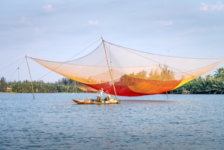 A fisher stands in a small boat on a calm body of water, operating a large, colorful fishing net suspended by poles. The net has a gradient of orange and yellow hues and stretches across the scene. Dense greenery lines the far shore under a blue sky scattered with a few clouds.