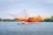 A fisher stands in a small boat on a calm body of water, operating a large, colorful fishing net suspended by poles. The net has a gradient of orange and yellow hues and stretches across the scene. Dense greenery lines the far shore under a blue sky scattered with a few clouds.
