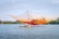 A fisher stands in a small boat on a calm body of water, operating a large, colorful fishing net suspended by poles. The net has a gradient of orange and yellow hues and stretches across the scene. Dense greenery lines the far shore under a blue sky scattered with a few clouds.