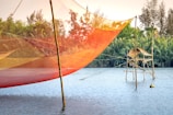 A vibrant fishing net hangs above a calm body of water, supported by wooden poles. In the background, lush greenery and trees enhance the scenic view, with a traditional bamboo structure present over the water's surface. The sky is clear, suggesting a peaceful, serene environment.