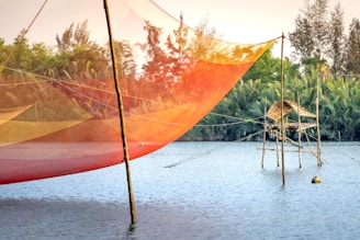 A vibrant fishing net hangs above a calm body of water, supported by wooden poles. In the background, lush greenery and trees enhance the scenic view, with a traditional bamboo structure present over the water's surface. The sky is clear, suggesting a peaceful, serene environment.