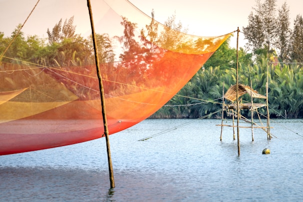 A vibrant fishing net hangs above a calm body of water, supported by wooden poles. In the background, lush greenery and trees enhance the scenic view, with a traditional bamboo structure present over the water's surface. The sky is clear, suggesting a peaceful, serene environment.