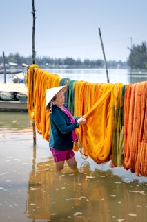 A person stands knee-deep in water, working with brightly colored fishing nets hanging on poles. The nets are mainly orange, yellow, and green. In the background, there is a blurred view of trees and structures across a body of water. The person is wearing a conical hat and casual clothing.