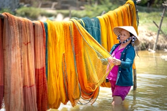 A person wearing a hat is working with colorful fishing nets hung to dry. The bright nets, including shades of orange, yellow, and teal, are draped over wooden poles above shallow water. The scene is outdoors and suggests a rural setting.