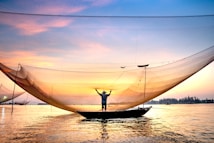 A fisherman stands on a small boat on a calm river, holding up a large fishing net against the backdrop of a vibrant sunrise or sunset. The sky is filled with hues of orange and pink, and the water reflects these colors, creating a serene and picturesque scene.