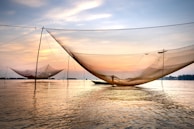 Large fishing nets are suspended above a calm body of water, with a boat in the distance and an orange-hued sunset in the sky. The scene conveys a peaceful and serene atmosphere, reflecting the beauty of nature and traditional fishing practices.