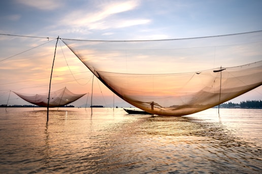 Large fishing nets are suspended above a calm body of water, with a boat in the distance and an orange-hued sunset in the sky. The scene conveys a peaceful and serene atmosphere, reflecting the beauty of nature and traditional fishing practices.