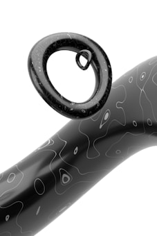 A minimalistic black and white 3D rendering featuring a smooth, curved torus shape suspended above a wavy cylindrical form. Both objects are characterized by a glossy finish with irregular line patterns that resemble contour lines on a map.