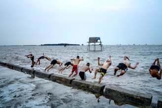A group of people in swimwear are performing synchronized backflips off a concrete barrier into the sea. The water is choppy and extends to the horizon. In the distance, a structure on stilts stands in the water, with more similar structures scattered across the horizon under a cloudy sky.