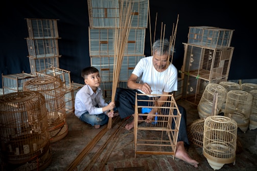 Two individuals are engaged in crafting birdcages. An older man is focused on assembling a wooden frame, while a young boy watches attentively. Various birdcages, both completed and in progress, surround them on the floor, hinting at a workshop setting.
