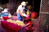 Two people are engaged in the process of making incense sticks. They are handling bundles of incense with bright pink and yellow colors. In the background, there are bags likely containing materials for the incense and large red pots placed on open fires, suggesting a traditional method of production. The setting seems to be a workshop with metal netting and fans visible.