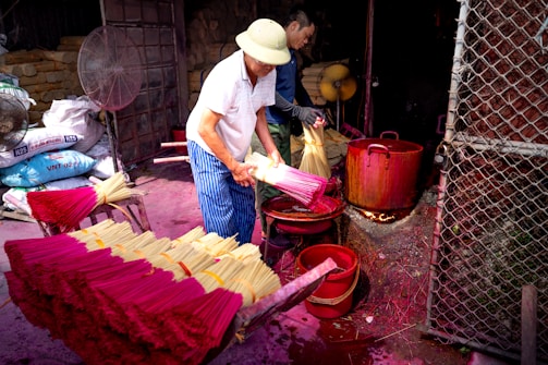 Two people are engaged in the process of making incense sticks. They are handling bundles of incense with bright pink and yellow colors. In the background, there are bags likely containing materials for the incense and large red pots placed on open fires, suggesting a traditional method of production. The setting seems to be a workshop with metal netting and fans visible.