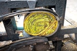 Close-up view of a machine with a roll of coiled material inside. The material appears to be layered with vibrant yellow and green tones, possibly depicting a leafy texture. The machine's metallic frame is dark and industrial, showing signs of wear and rust.