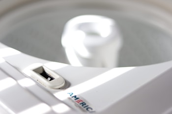 A close-up of a white washing machine, focusing on the top area with a dial and the inner tub visible. Sunlight casts soft stripes across the surface, creating a serene atmosphere.