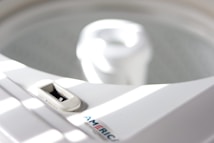 A close-up of a white washing machine, focusing on the top area with a dial and the inner tub visible. Sunlight casts soft stripes across the surface, creating a serene atmosphere.