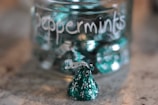 A glass jar filled with green-wrapped peppermint candies sits on a surface. The jar is labeled 'Peppermints' in white handwriting. One candy is outside the jar, prominently in the foreground.