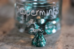 A glass jar filled with green-wrapped peppermint candies sits on a surface. The jar is labeled 'Peppermints' in white handwriting. One candy is outside the jar, prominently in the foreground.