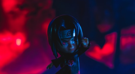 A small figurine of a character with long dark hair and a headband featuring a symbol is positioned prominently. The background is filled with intense red and blue coloring, creating a dramatic and moody atmosphere.