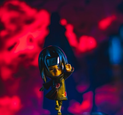 A stylized figurine of a character with intense expression, dressed in a dark cloak with red cloud patterns, set against a vibrant red and dark blue background. The character is depicted in a dynamic pose, suggesting motion or action.