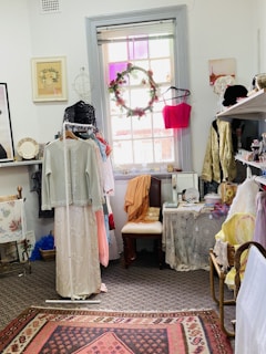 A small room decorated with vintage clothing and accessories. A dress on a hanger is displayed prominently in front of a window, which has a colorful wreath hanging on it. Shelves are lined with clothes, hats, and small decorative items. A chair covered with a lace throw sits beneath the window, surrounded by other vintage-style decor.