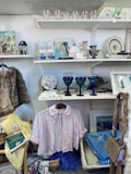 A variety of vintage and decorative items are displayed on shelves and hanging. On the shelves, there are glassware items, framed pictures, and a teapot. A hanging pink blouse is centrally featured, surrounded by baskets, scarves, and books. The overall appearance is reminiscent of a thrift or antique shop.