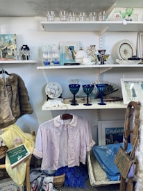 A variety of vintage and decorative items are displayed on shelves and hanging. On the shelves, there are glassware items, framed pictures, and a teapot. A hanging pink blouse is centrally featured, surrounded by baskets, scarves, and books. The overall appearance is reminiscent of a thrift or antique shop.
