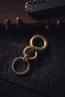 A vintage-style notebook with leather binding and visible stitching lies on a dark surface. Resting on it is a multi-tool featuring a corkscrew, alongside a brass key ring consisting of interconnected rings with engraved texts.