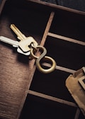 A set of keys attached to a brass keyring with intricate circular designs are placed inside a wooden compartmentalized organizer. There is also a piece of brown paper or cardboard partially visible in the bottom right corner.