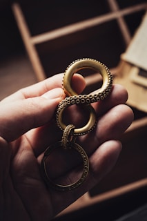 A hand holds a gold-colored metal object shaped like the number eight with textured details, attached to a circular ring. The background features blurred wooden compartments.