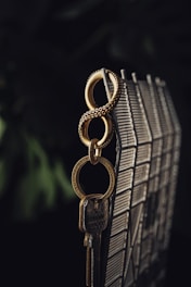 A gold-colored keychain with intricate detailing hangs from a metallic grid. The keychain holds a set of keys, casting a soft shadow against the dark background. The lighting emphasizes the textures and patterns on the keychain and the keys.
