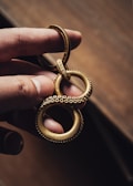 A hand holds a gold-colored metallic keychain shaped like an infinity symbol with a textured, beaded surface, resembling tentacles. The background is a blurred, warm-toned surface, emphasizing the intricate design and craftsmanship of the keychain.