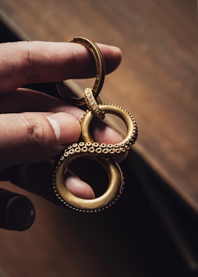 A hand holds a gold-colored metallic keychain shaped like an infinity symbol with a textured, beaded surface, resembling tentacles. The background is a blurred, warm-toned surface, emphasizing the intricate design and craftsmanship of the keychain.