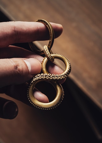 A hand holds a gold-colored metallic keychain shaped like an infinity symbol with a textured, beaded surface, resembling tentacles. The background is a blurred, warm-toned surface, emphasizing the intricate design and craftsmanship of the keychain.