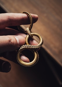 A hand holds a gold-colored metallic keychain shaped like an infinity symbol with a textured, beaded surface, resembling tentacles. The background is a blurred, warm-toned surface, emphasizing the intricate design and craftsmanship of the keychain.