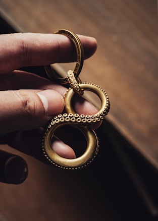 A hand holds a gold-colored metallic keychain shaped like an infinity symbol with a textured, beaded surface, resembling tentacles. The background is a blurred, warm-toned surface, emphasizing the intricate design and craftsmanship of the keychain.