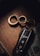 A luxury car key fob is placed on a wooden surface alongside a set of metallic rings in a figure-eight formation. Partially visible underneath is a piece of currency, suggesting a sense of affluence.