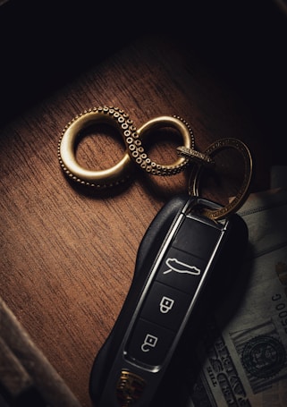 A luxury car key fob is placed on a wooden surface alongside a set of metallic rings in a figure-eight formation. Partially visible underneath is a piece of currency, suggesting a sense of affluence.