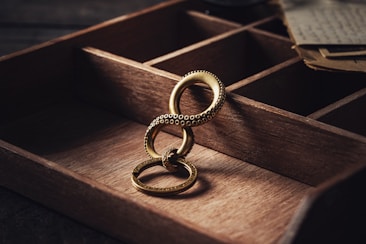 A gold ring with an intricate design featuring overlapping circular patterns lies in a wooden compartmentalized box. The background includes a few pieces of vintage paper, adding a rustic and historical ambiance.