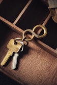 A close-up view of a set of keys attached to a unique keyring with an intertwined double ring design. The keys and keyring are placed on the edge of a wooden compartmentalized organizer, with different sections visible and a piece of torn paper partially seen in the upper right corner.