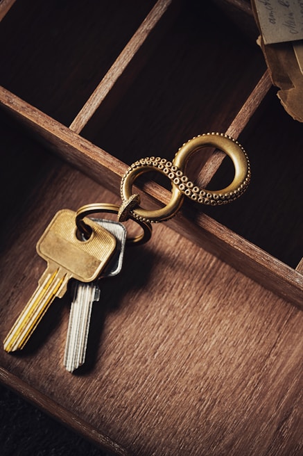 A close-up view of a set of keys attached to a unique keyring with an intertwined double ring design. The keys and keyring are placed on the edge of a wooden compartmentalized organizer, with different sections visible and a piece of torn paper partially seen in the upper right corner.