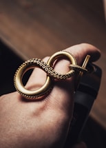 A hand is adorned with a large, intricately designed gold ring shaped like an infinity symbol. The ring features small, detailed dot patterns around its edges, evoking a sense of luxury and craftsmanship. The lighting accentuates the gold's rich finish against the skin tone.