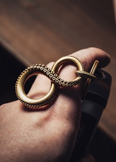 A hand is adorned with a large, intricately designed gold ring shaped like an infinity symbol. The ring features small, detailed dot patterns around its edges, evoking a sense of luxury and craftsmanship. The lighting accentuates the gold's rich finish against the skin tone.