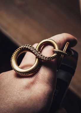 A hand is adorned with a large, intricately designed gold ring shaped like an infinity symbol. The ring features small, detailed dot patterns around its edges, evoking a sense of luxury and craftsmanship. The lighting accentuates the gold's rich finish against the skin tone.