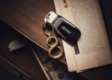 A black car key with a metallic keychain featuring three interconnected loops lies on an open newspaper. The background consists of a wooden surface with a folded knife and a partially visible banknote inside a drawer.