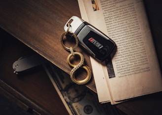 A black car key with a metallic keychain featuring three interconnected loops lies on an open newspaper. The background consists of a wooden surface with a folded knife and a partially visible banknote inside a drawer.