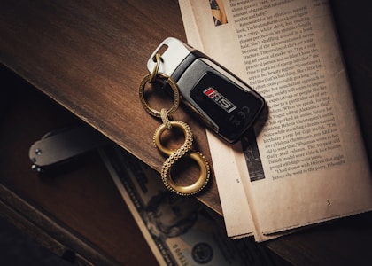A black car key with a metallic keychain featuring three interconnected loops lies on an open newspaper. The background consists of a wooden surface with a folded knife and a partially visible banknote inside a drawer.