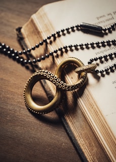 A close-up of an old book with worn pages, partially open, lying on a wooden surface. A decorative, antique-looking keychain with an infinity symbol made of textured metal is placed on top, alongside a string of dark beads resembling a rosary or necklace.