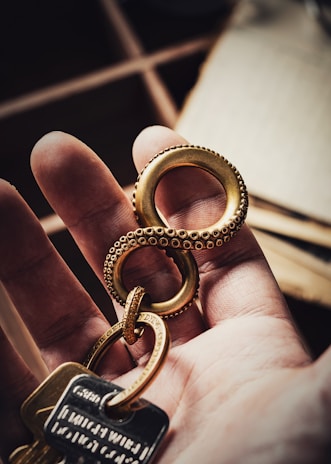 A hand is holding a gold-colored keychain shaped like an infinity symbol with a textured design resembling tentacles. Several keys are attached to the keychain, including one with an engraved tag. The background is softly blurred, focusing attention on the hand and keychain.