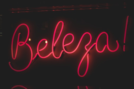 Bright pink neon sign displaying the word 'Beleza' against a dark background. The glowing letters have a cursive style, creating an eye-catching and lively appearance.